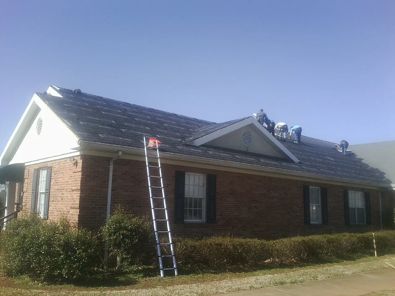 Crew tearing shingles off a brick ranch roof with a ladder set — Emergency Roof Repair work by our crew in Fort Hill