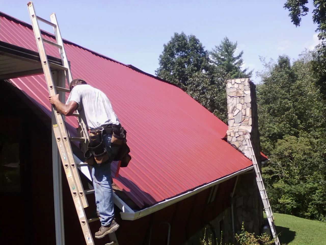 Roofer on a ladder beside a red metal roof and stone chimney — Soffit & Fascia Repair by Roofing Brothers in Fort Hill